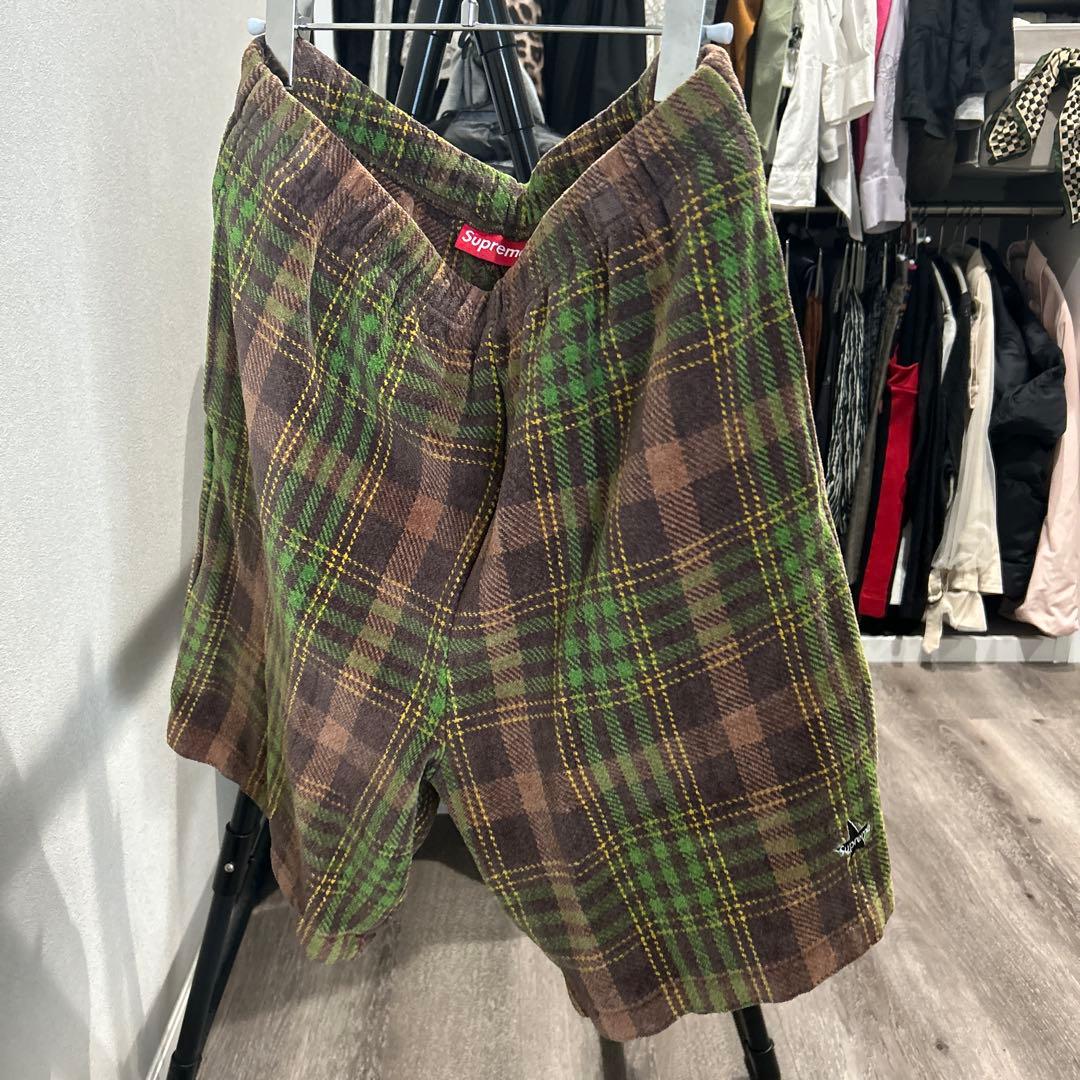 Brown and green plaid velour short pants with a red Supreme label hanging on a hanger against a closet background.