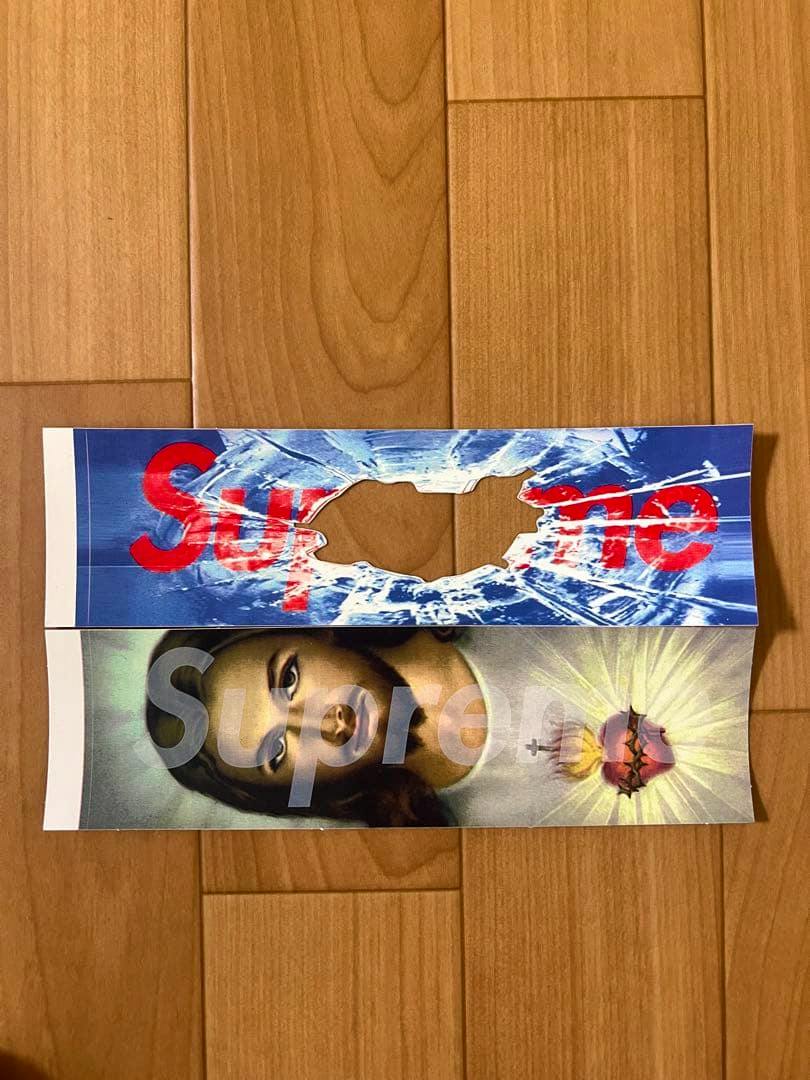 Two Supreme stickers laid flat on a wooden floor: a blue cracked-ice design with a central tear and a Jesus portrait sticker with a translucent "Supreme" logo over a sacred heart.