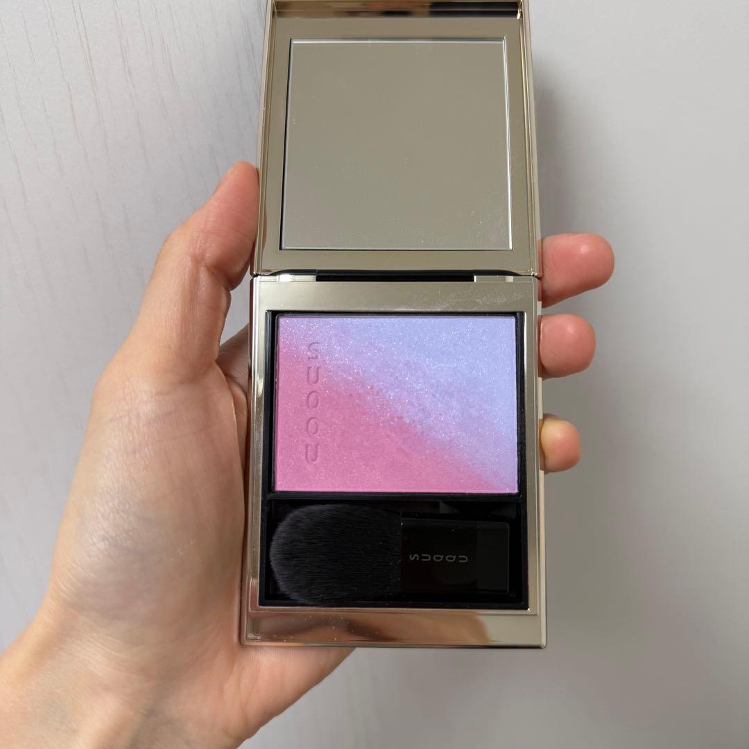 Hand holding an open SUQQU Blurring Color Blush compact (shade 113) showing a pink-to-lavender gradient powder, built-in mirror, and included brush.