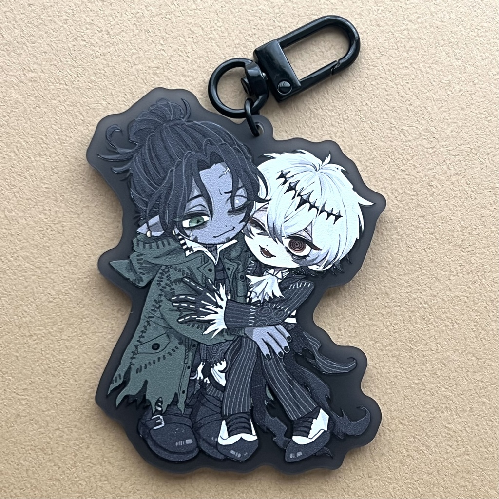 Rubber keychain showing two chibi characters embracing, attached to a black clasp on a beige background.