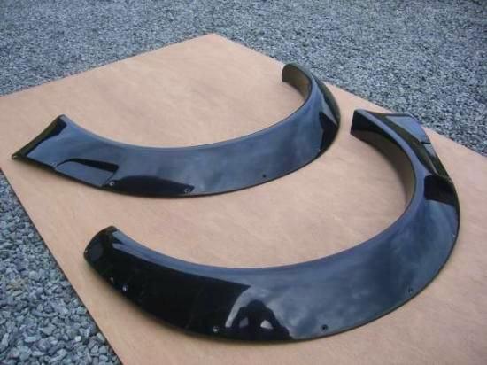 Two black FRP 50mm wide rear fender flares for a Cappuccino displayed on cardboard.