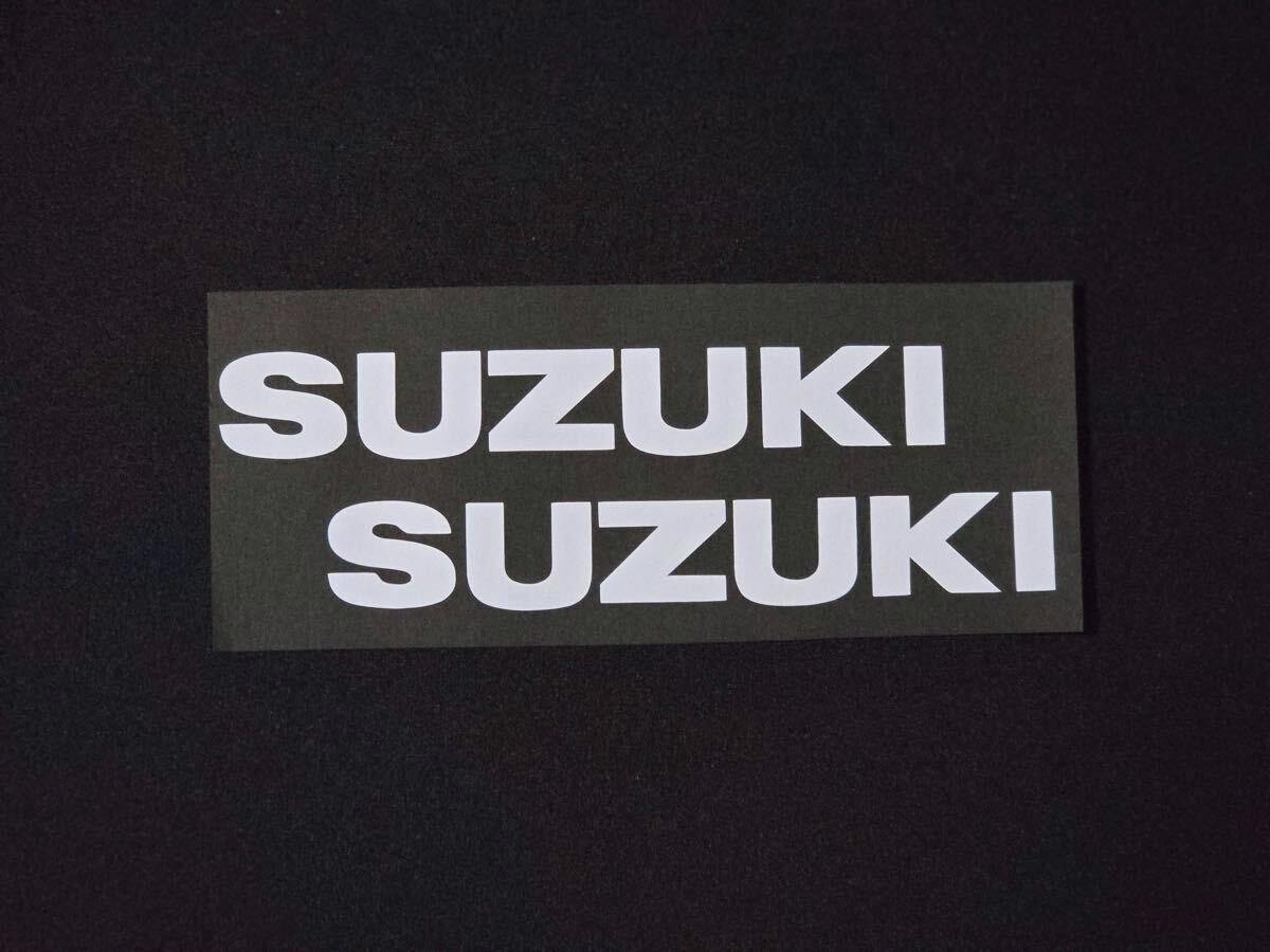 Two large white "SUZUKI" car window stickers on a dark backing sheet against a dark background.