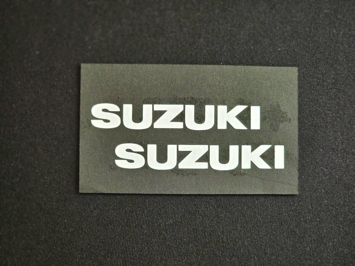 Two white "SUZUKI" text car stickers on a rectangular black backing placed on a dark surface.
