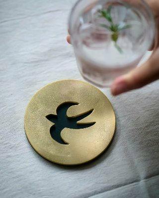 Round metal coaster with a swallow-shaped cutout on a fabric surface, a hand holding a glass visible above it.