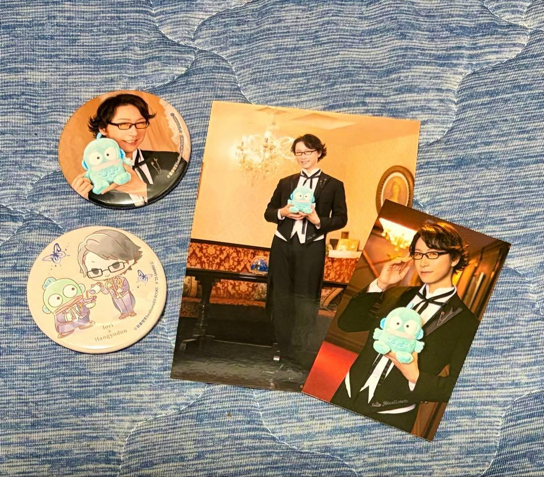 Two round can badges and two photo cards showing 伊織 in a butler outfit holding a blue Sanrio character, displayed on a blue fabric background.