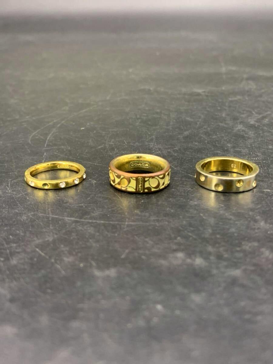 Three gold-tone rings—center ring with Coach signature pattern—placed side-by-side on a dark scratched surface.