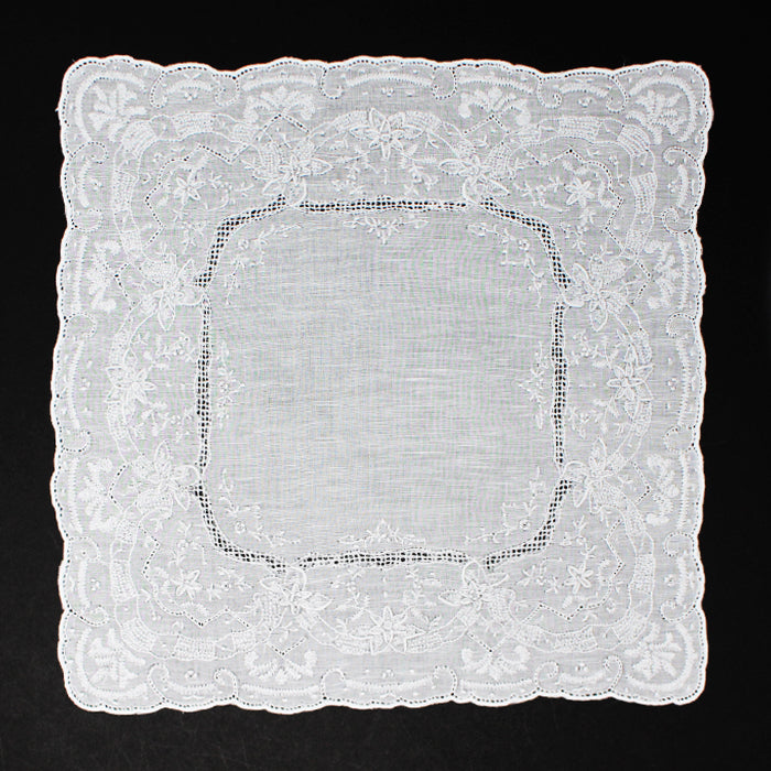 A white hand-embroidered handkerchief with a scalloped lace border and floral embroidery laid flat on a black background.