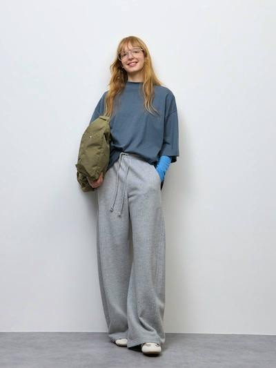 Model wearing light gray sweat barrel pants paired with a blue oversized top and holding an olive jacket.