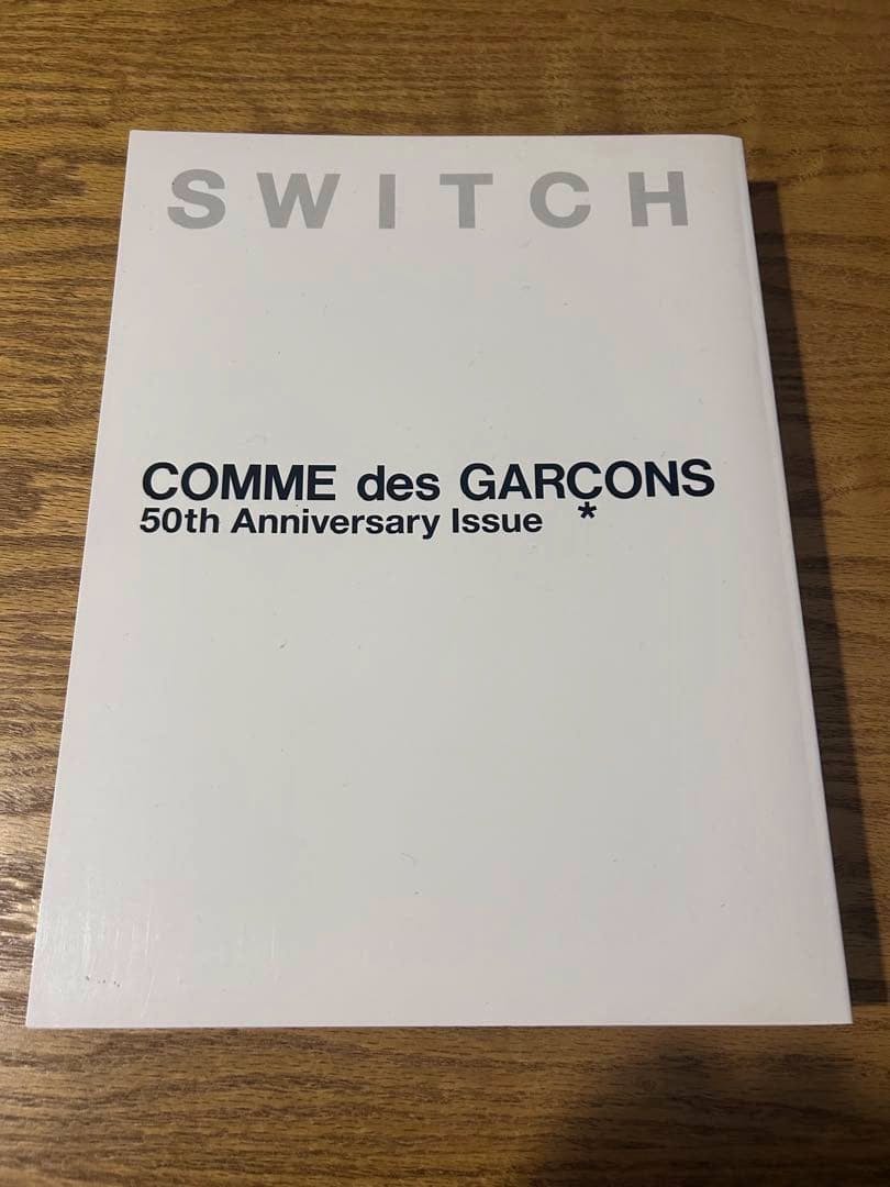 Front cover of the SWITCH COMME des GARCONS 50thAnniversary magazine with title text on a white cover placed on a wooden surface.
