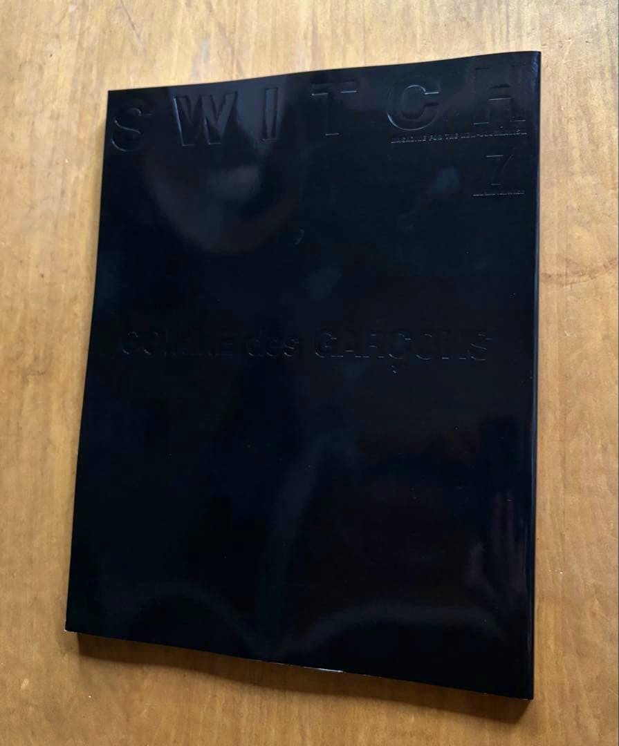 A black glossy magazine (SWITCH) with embossed "SWITCH" and "COMME des GARÇONS" lettering lying on a wooden surface.