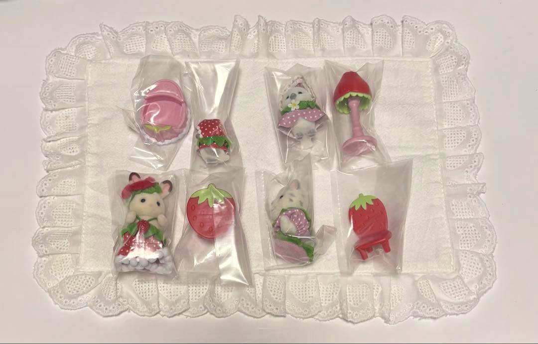 Sylvanian Families baby furniture and small animal figures with strawberry motifs, each in clear plastic bags, arranged on a white lace mat.