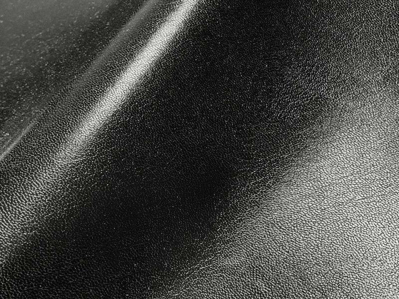 Close-up of thin black glossy synthetic leather (polyester) fabric showing its pebbled texture and light reflections.