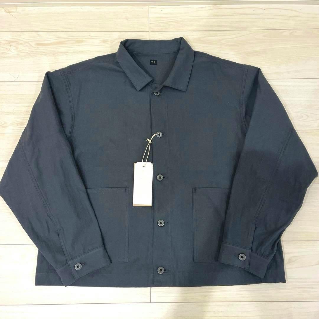 A dark cotton men's coverall laid flat on a light wood floor showing front buttons, collar, two patch pockets, and a white hang tag.