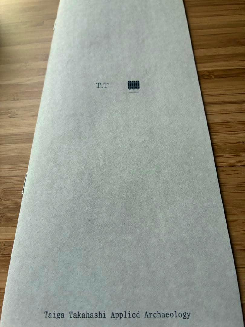Front of the pamphlet titled "Taiga Takahashi Applied Archaeology" on textured light gray paper with "T.T" and a small logo, photographed on a wooden surface.