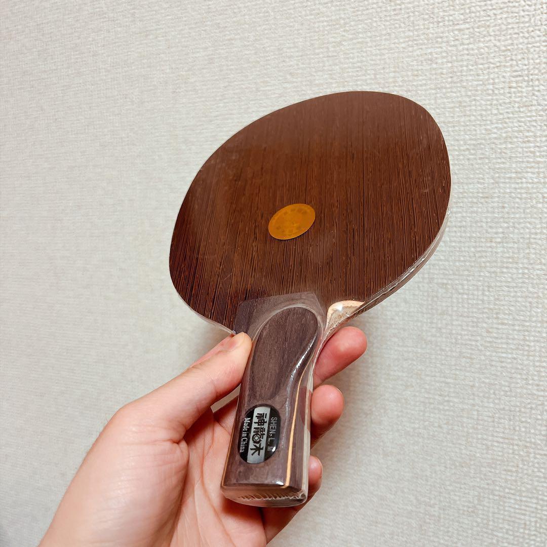Hand holding a Chinese-style penhold table tennis racket with a dark wooden blade against a light wall.