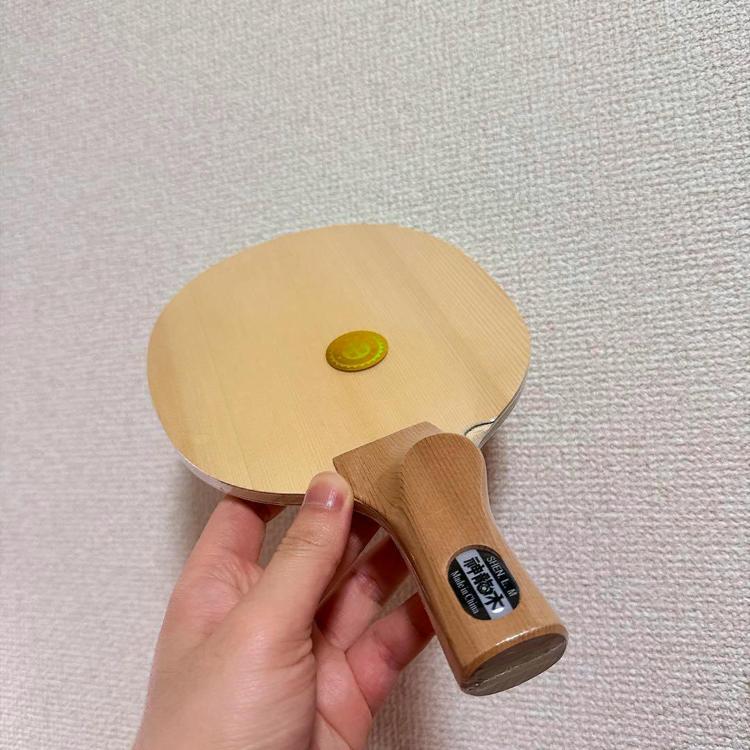 A hand holds the table tennis racket (Chinese-style penhold blade) showing the wooden handle and bare blade surface against a textured wall.