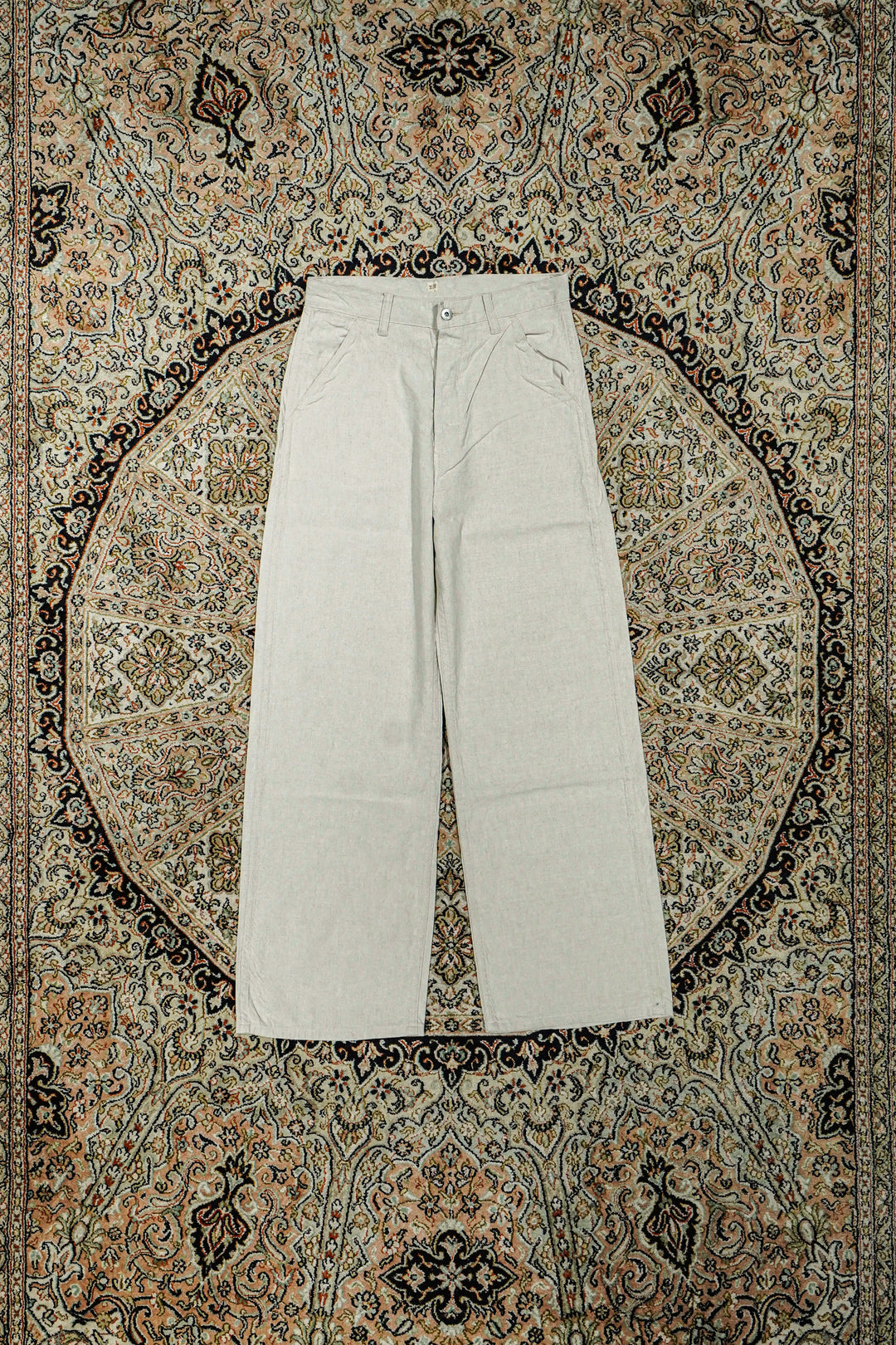 Flat lay of Taiga Takahashi women's ivory linen painter pants laid flat on a patterned rug.