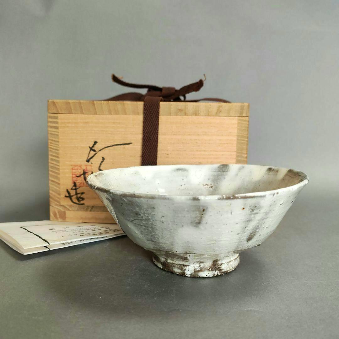 A white-glazed ceramic tea bowl (chawan) shown with its wooden storage box and paperwork behind it.