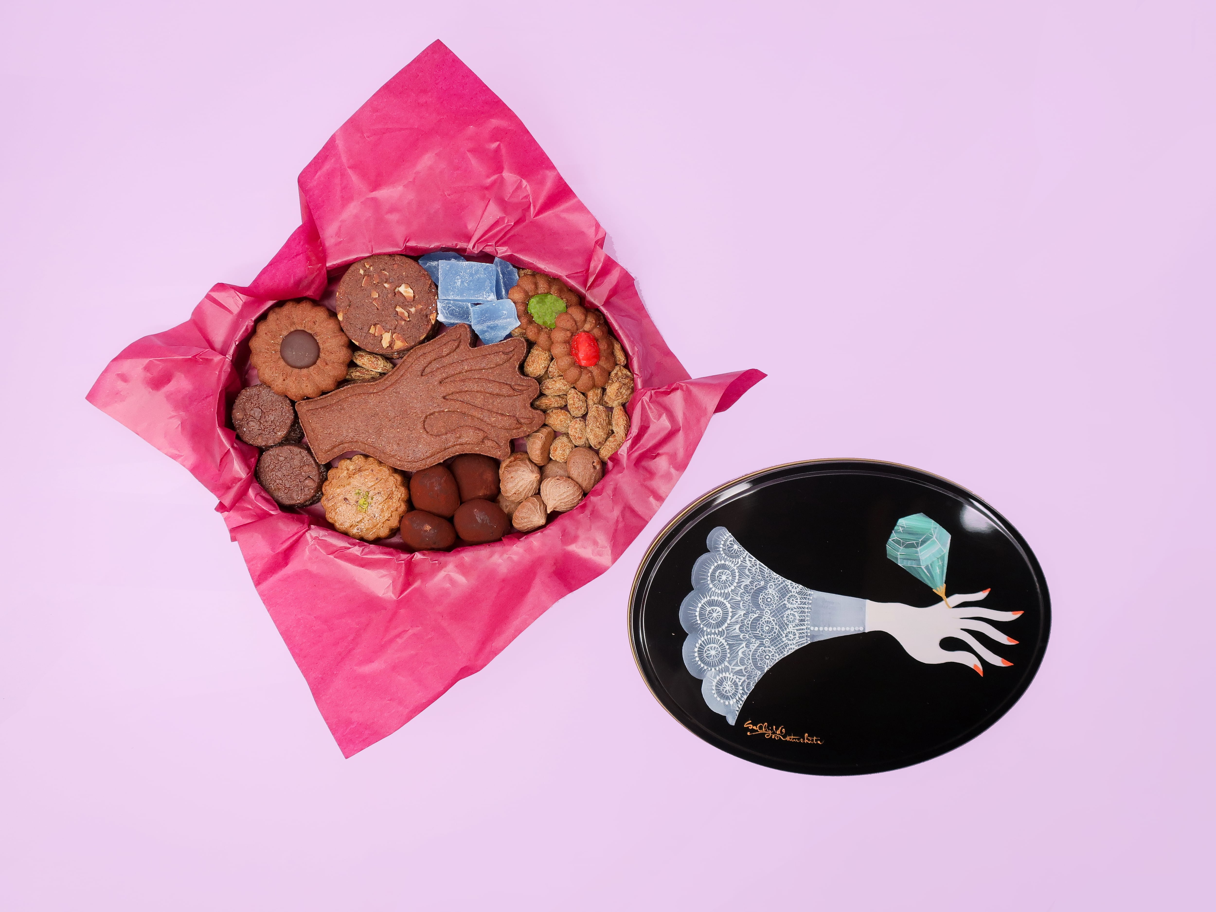 An open oval tin of packed assorted cookies with the tin lid beside it on a pink background.