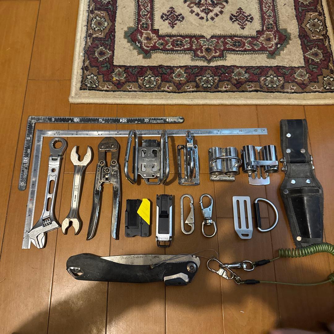 An array of metal hand tools laid out on a wooden floor, including an adjustable wrench, open-end wrenches, pliers, rulers, clips, and a tool holster.