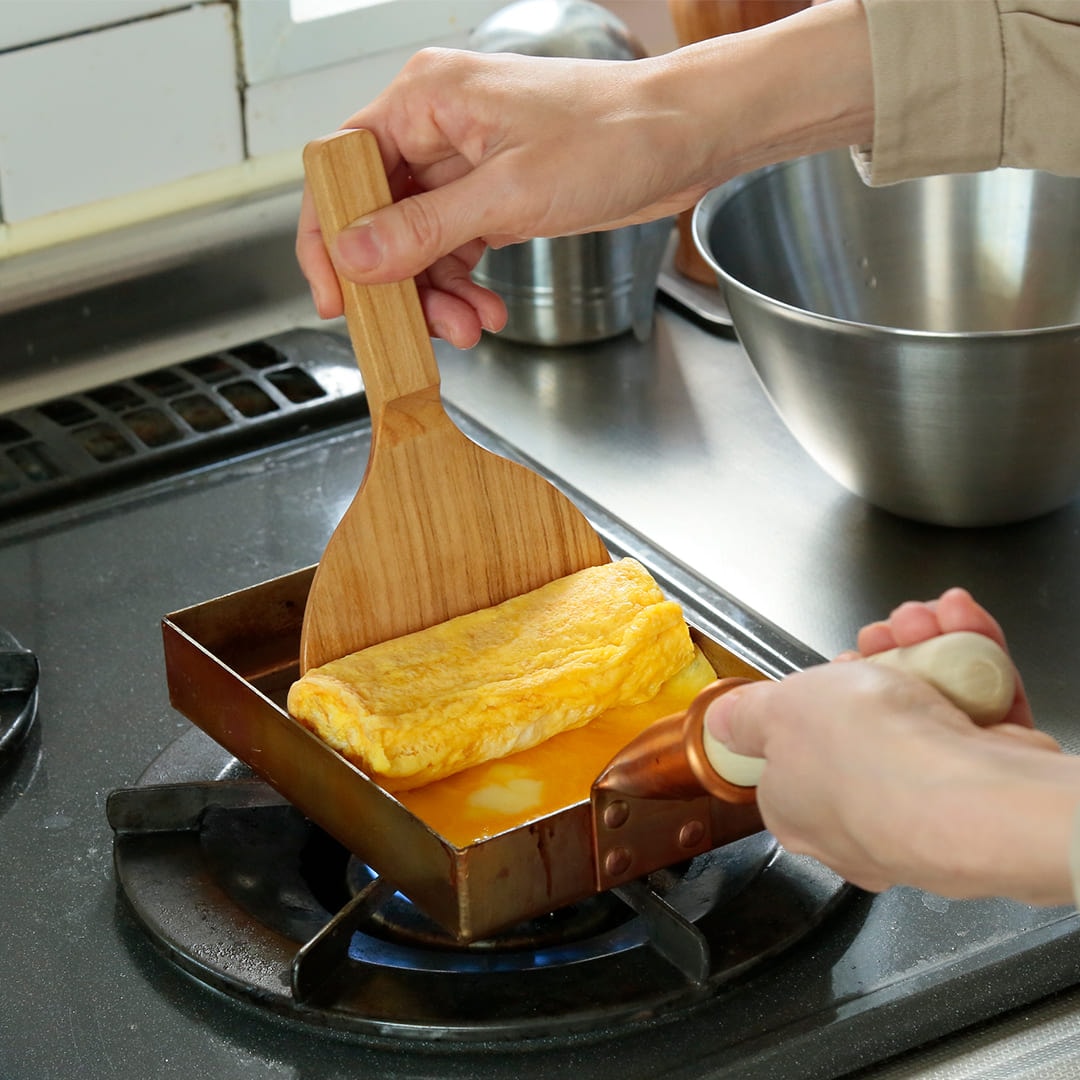 A hand using a wooden tamagoyaki spatula to roll an omelette in a rectangular pan on a stovetop.