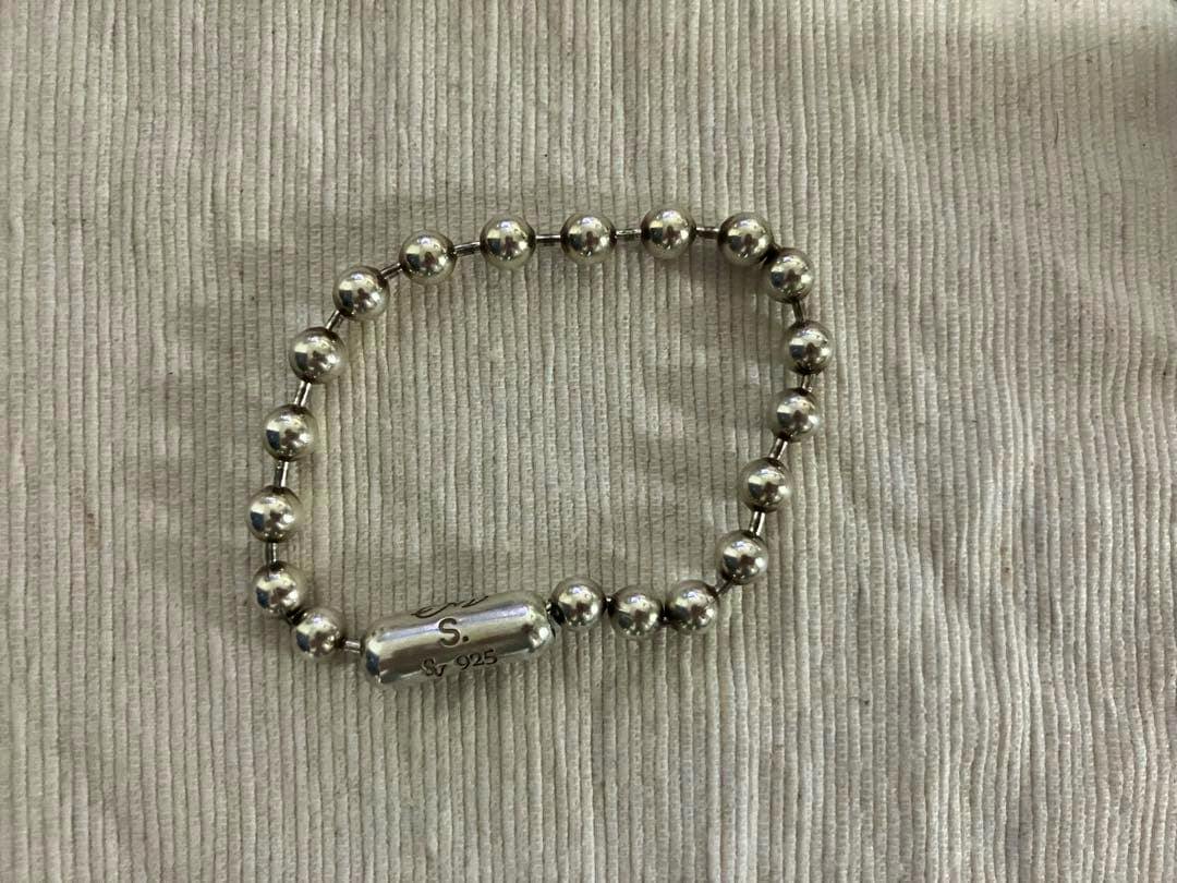A silver ball-chain bracelet with a cylindrical clasp stamped "S" and "925" laid on a ribbed fabric background.