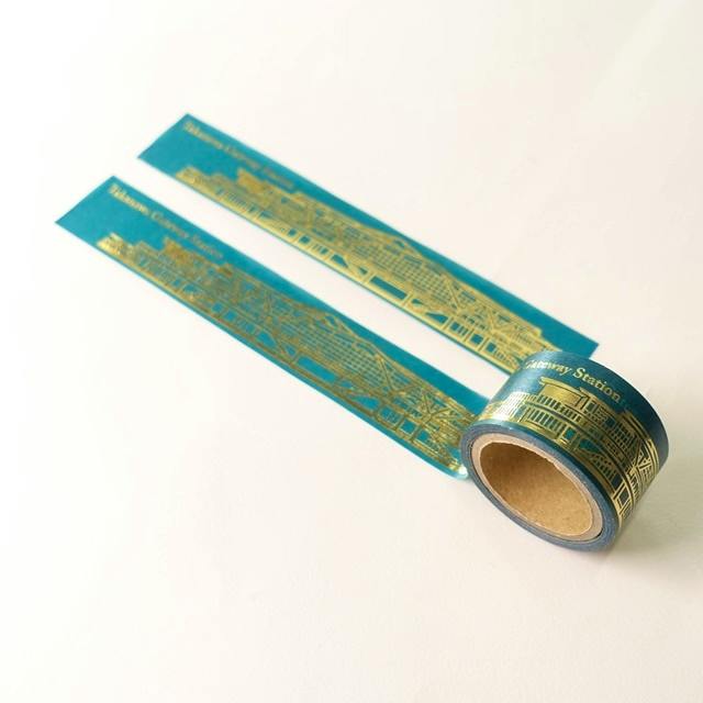 A teal masking tape roll printed with a gold illustration of Takanawa Gateway Station with two strips of tape unrolled on a white background.