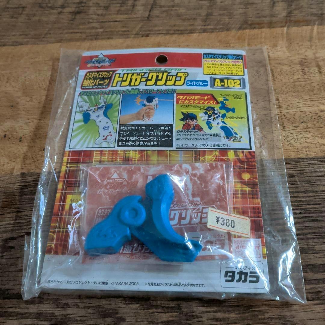 Light-blue Takara Beyblade A-102 trigger grip plastic part shown in its original blister packaging on a wooden surface.