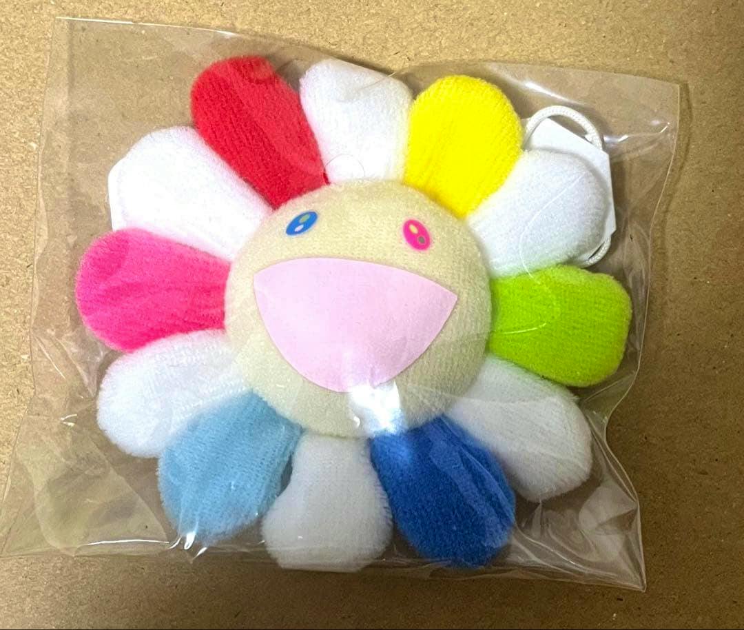 Plush rainbow flower badge/strap inside clear plastic packaging, showing multicolored petals and a smiling face.