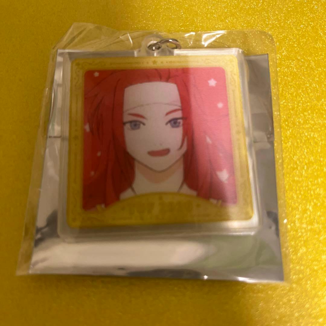 Sealed acrylic keychain in its packaging showing a square illustration of a red-haired character against a pink background on a yellow surface.