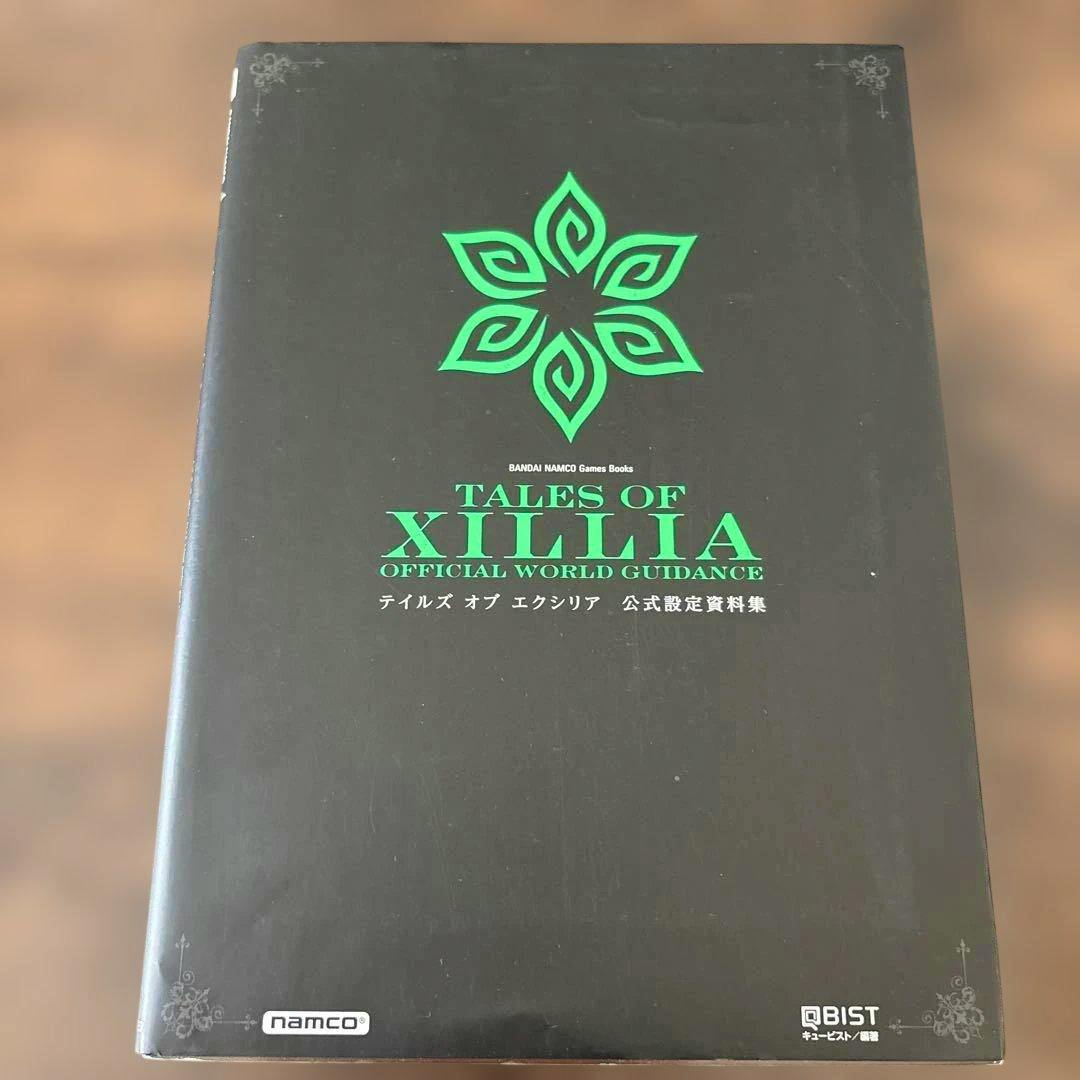 Front cover of a black Tales of Xillia official art book showing a green emblem and the text "TALES OF XILLIA OFFICIAL WORLD GUIDANCE."