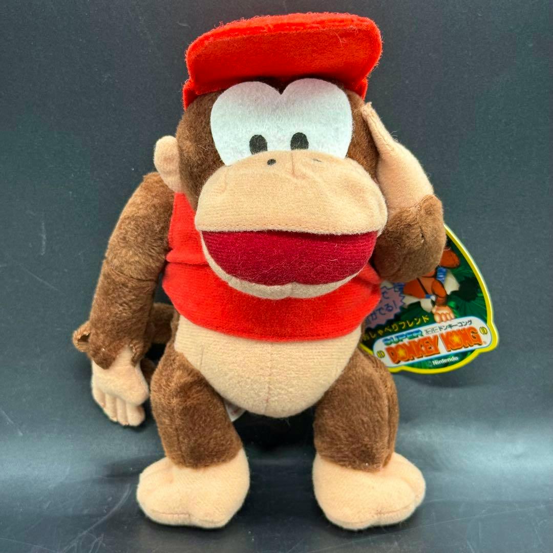 Front view of a Diddy Kong plush wearing a red hat and red shirt against a dark background.