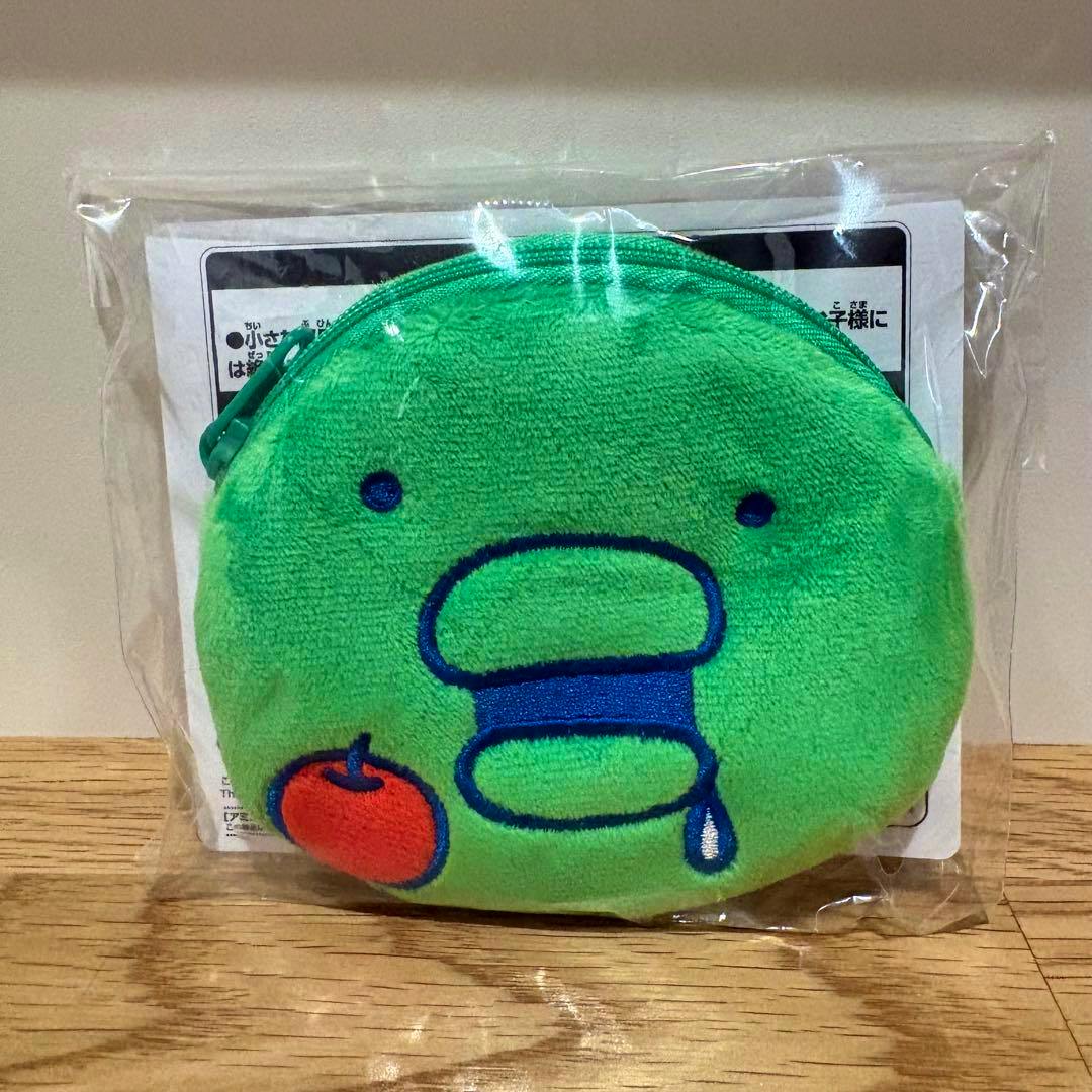 Green round Tamagotchi face pouch (Kuchipatchi) in clear plastic packaging showing embroidered eyes, mouth, and a small orange fruit motif.