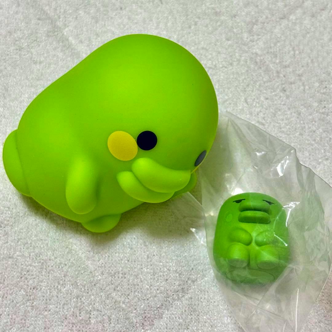 Photo of two green rubber Tamagotchi character toys, one larger figure and one smaller figure inside a clear plastic bag, on a light fabric surface.