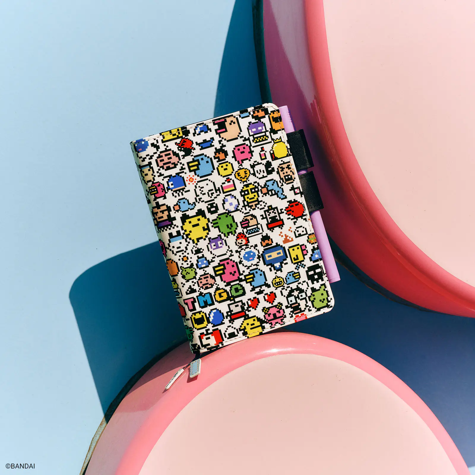 A small A6 book cover with colorful Tamagotchi pixel-art characters on the front, photographed on pink circular props against a blue background.