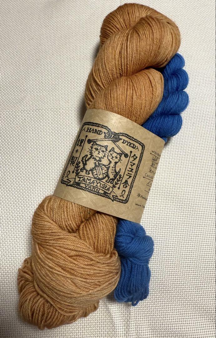 Two hand-dyed Tamayura yarn skeins, one brown-gold and one bright blue, with a Tamayura paper label on a light fabric background.