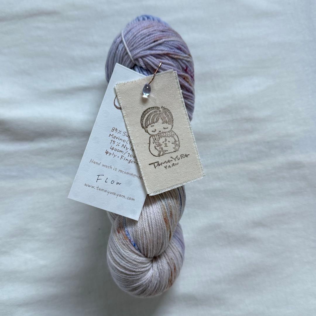 A hand-dyed Tamayura sock yarn skein lying on fabric with attached tags showing the brand illustration and fiber details.
