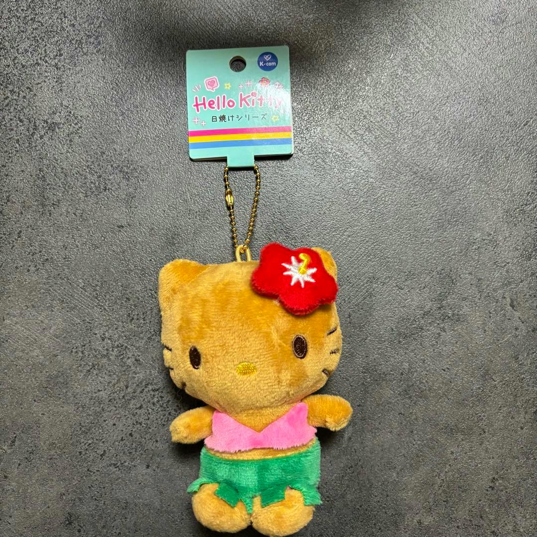 Front view of the Hello Kitty plush keychain showing a tan Hello Kitty wearing a pink top, green skirt, red flower, and attached retail tag.