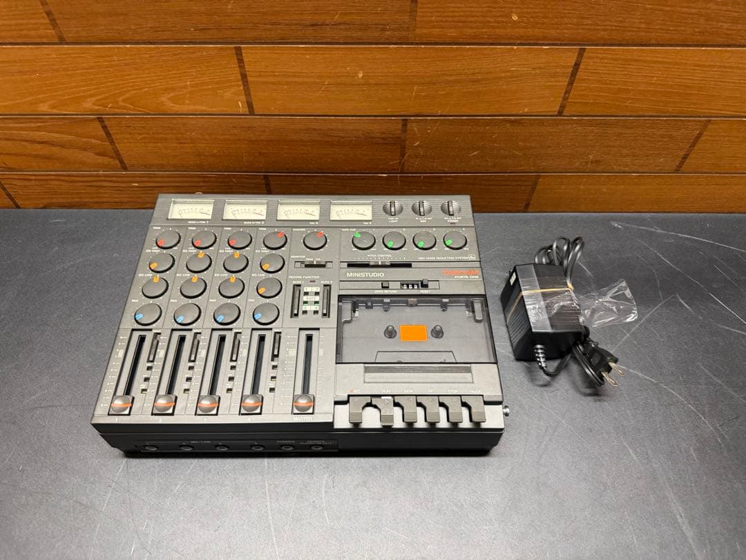 Front view of a TASCAM PORTA ONE ministudio cassette MTR on a black surface with its AC adapter beside it.
