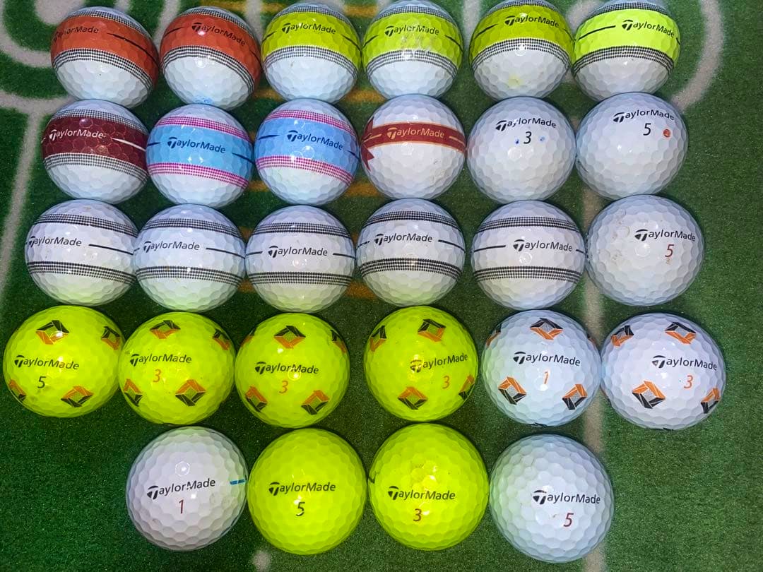 Twenty-eight TaylorMade golf balls arranged in rows on green turf, showing various colored markings.