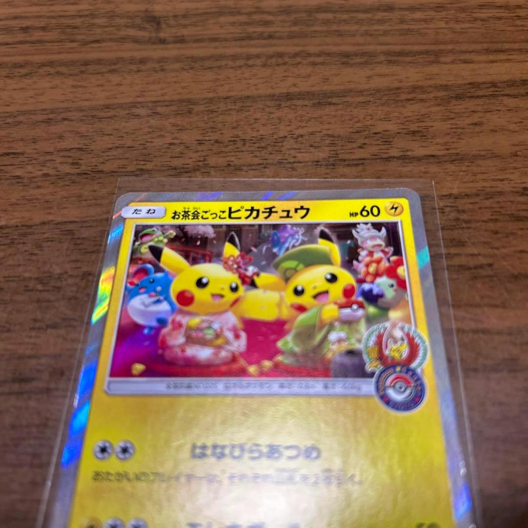 Photo of a holographic Pokémon trading card (お茶会ごっこピカチュウ) showing two Pikachu in tea-party outfits on a wood surface.
