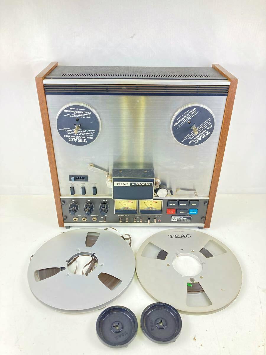 Front view of a TEAC A-3300SX open reel deck upright with two mounted reels and two empty reel hubs placed in front on a white background.