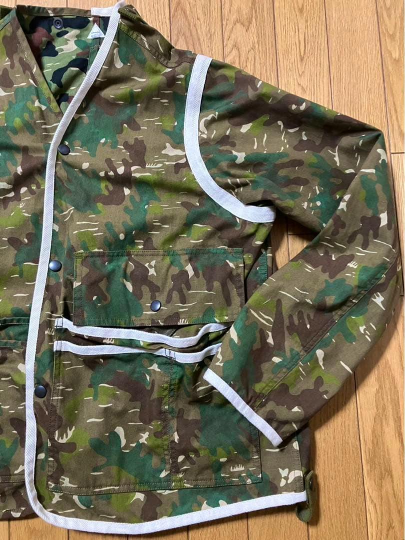 Front of a men's cotton camouflage jacket with white trim, flap pocket and snap buttons laid flat on a wooden floor.