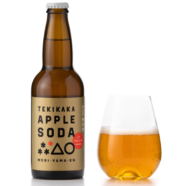 A brown glass bottle labeled "TEKIKAKA APPLE SODA" shown next to a stemless glass partially filled with apple-colored soda.
