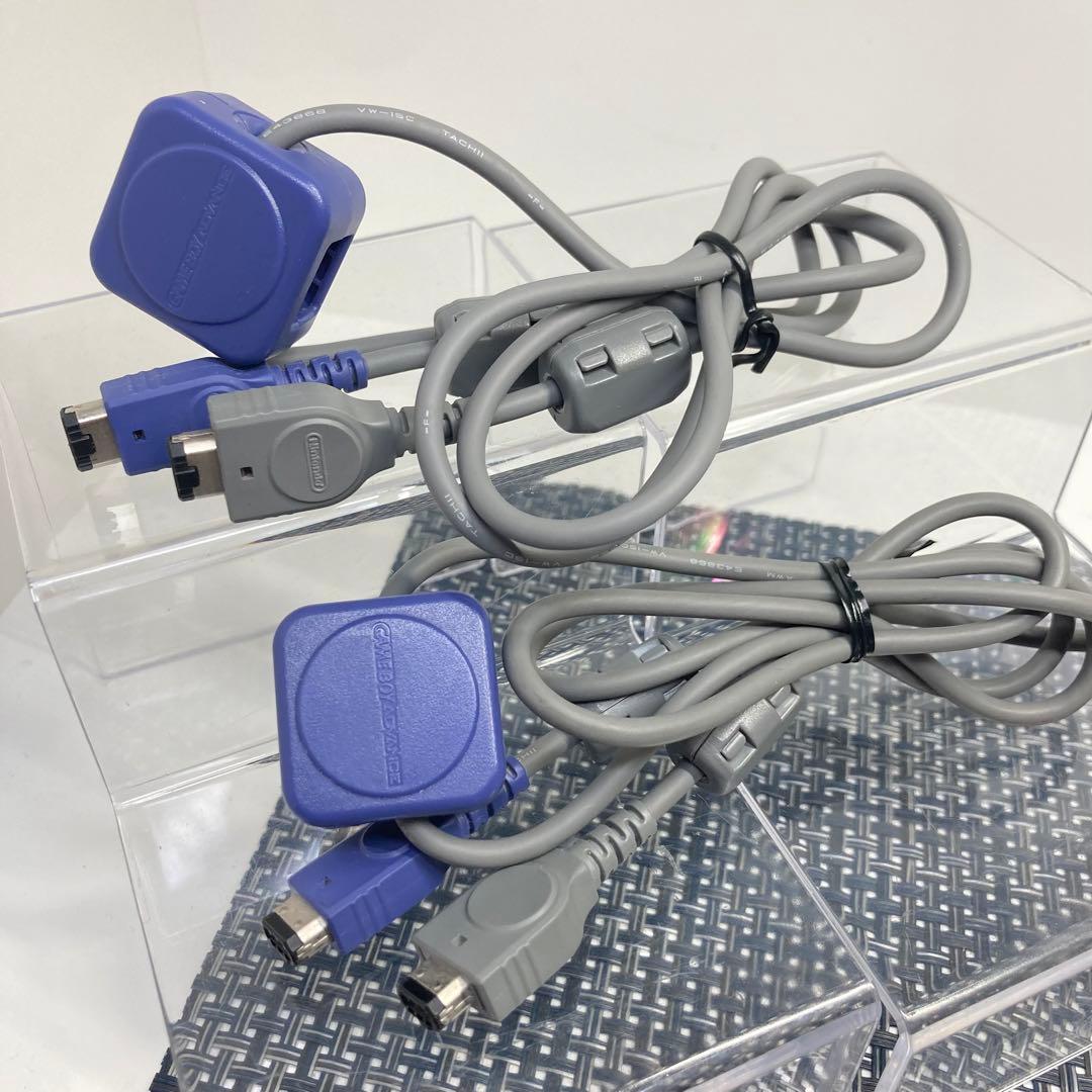 Two Game Boy Advance communication/link cables (two-piece set) with purple and gray connectors coiled on a clear display stand.