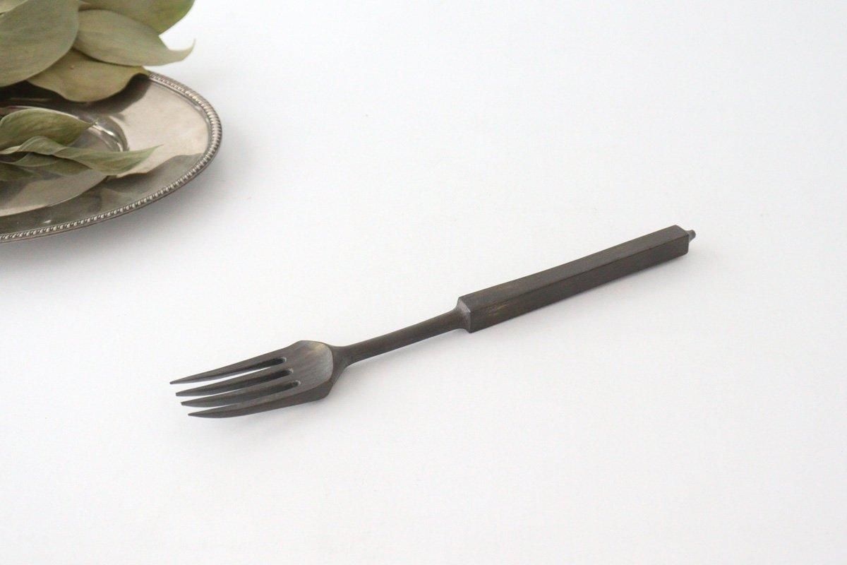 An antique metal fork with a rectangular handle shown on a white background beside a small plate and leaves.