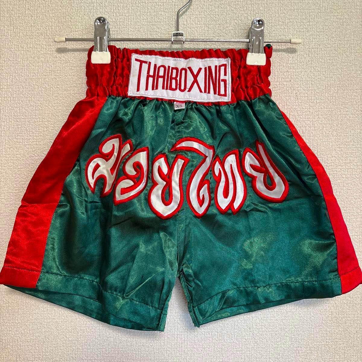 Green and red men's Muay Thai polyester shorts hanging on a hanger, showing a 'THAIBOXING' waist patch and white-and-red decorative Thai script on the front.