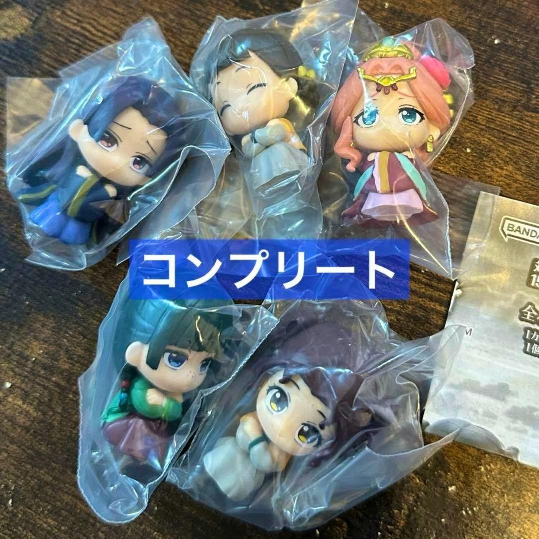 Five small sealed plastic chibi figures from the 薬屋のひとりごと series in clear bags arranged on a wooden surface with a blue “コンプリート” overlay.