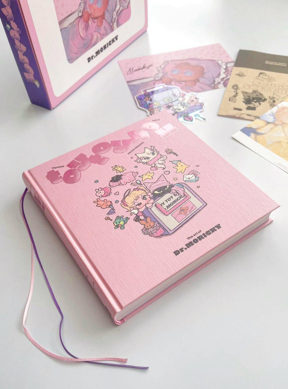 Pink hardcover book from "The Art of Dr.MORICKY" special edition shown on a white surface with ribbon bookmarks, with its box and inserts partially visible.