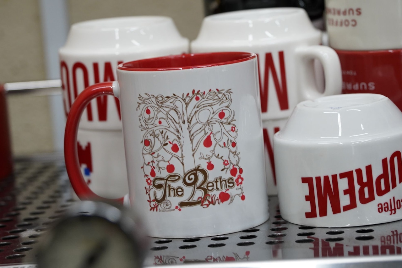 White ceramic two-tone mug with a red interior and handle, featuring The Beths tree logo on the front.